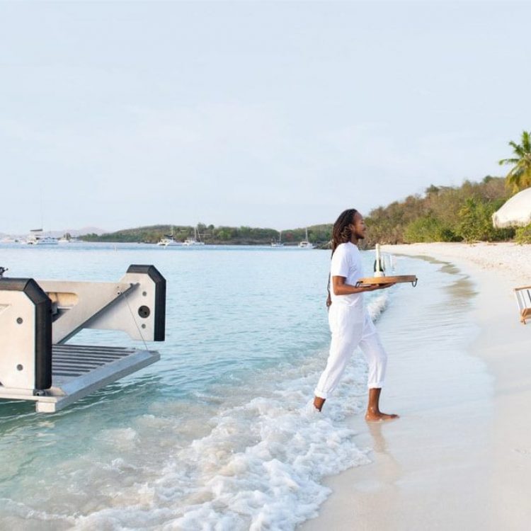 Resort staff member carrying champagne ashore from a boat on a white-sand Caribbean beach